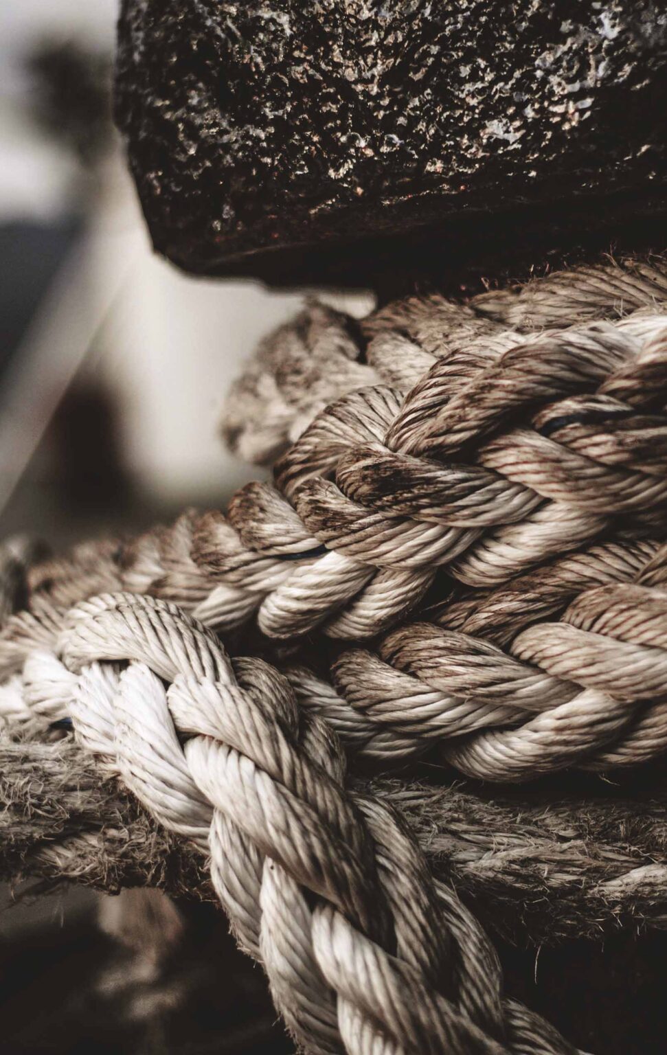 A close-up of a thick, twisted rope in muted beige tones, giving a sense of strength and durability. The texture of the rope is prominently displayed, with individual strands visibly interwoven. The background is out of focus, but one can discern what appears to be a weathered, rough surface, possibly metal, suggesting a maritime or industrial setting. The detailed focus on the rope highlights its robust structure and the essential role it plays in securing heavy objects, likely in a shipping or nautical context.