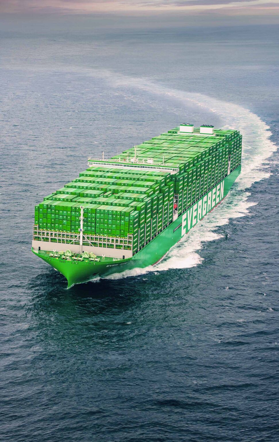 A large, green container ship named "EVERGREEN" is seen from a front-side angle as it navigates through the ocean. The ship's hull is loaded with rows of green containers, and it is cutting through the water, leaving a frothy white trail behind. The ocean around it is a deep blue and the sky above shows a soft gradient from gray to pale pink, possibly indicating early morning or late evening light.