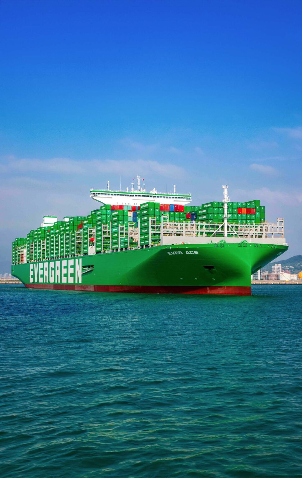 A large green Evergreen cargo ship loaded with numerous green containers is afloat on calm blue waters. The sky is clear with few clouds, and a faint coastline is visible in the background. The ship's hull is marked with the name "EVER AGE" in white letters.