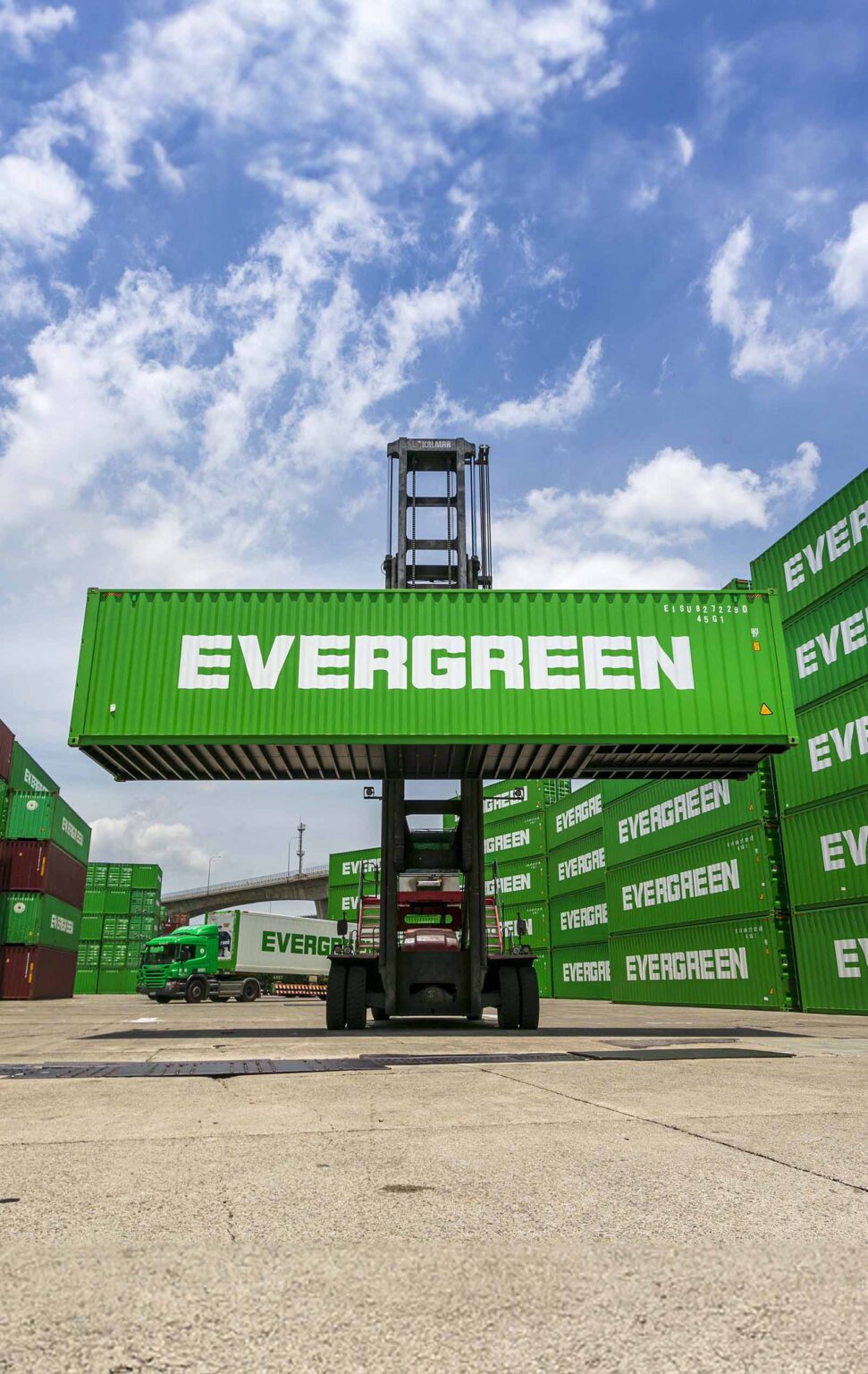 A straddle carrier is shown lifting a green container emblazoned with the white "EVERGREEN" logo. The machine is centered in the image, with a clear blue sky filled with white clouds overhead. In the background, there's a neatly arranged stack of similar green containers creating a vibrant, uniform backdrop. To the left, a green truck featuring the same logo is parked, adding to the busy logistics atmosphere of a container terminal. The concrete ground suggests a well-traveled industrial area.