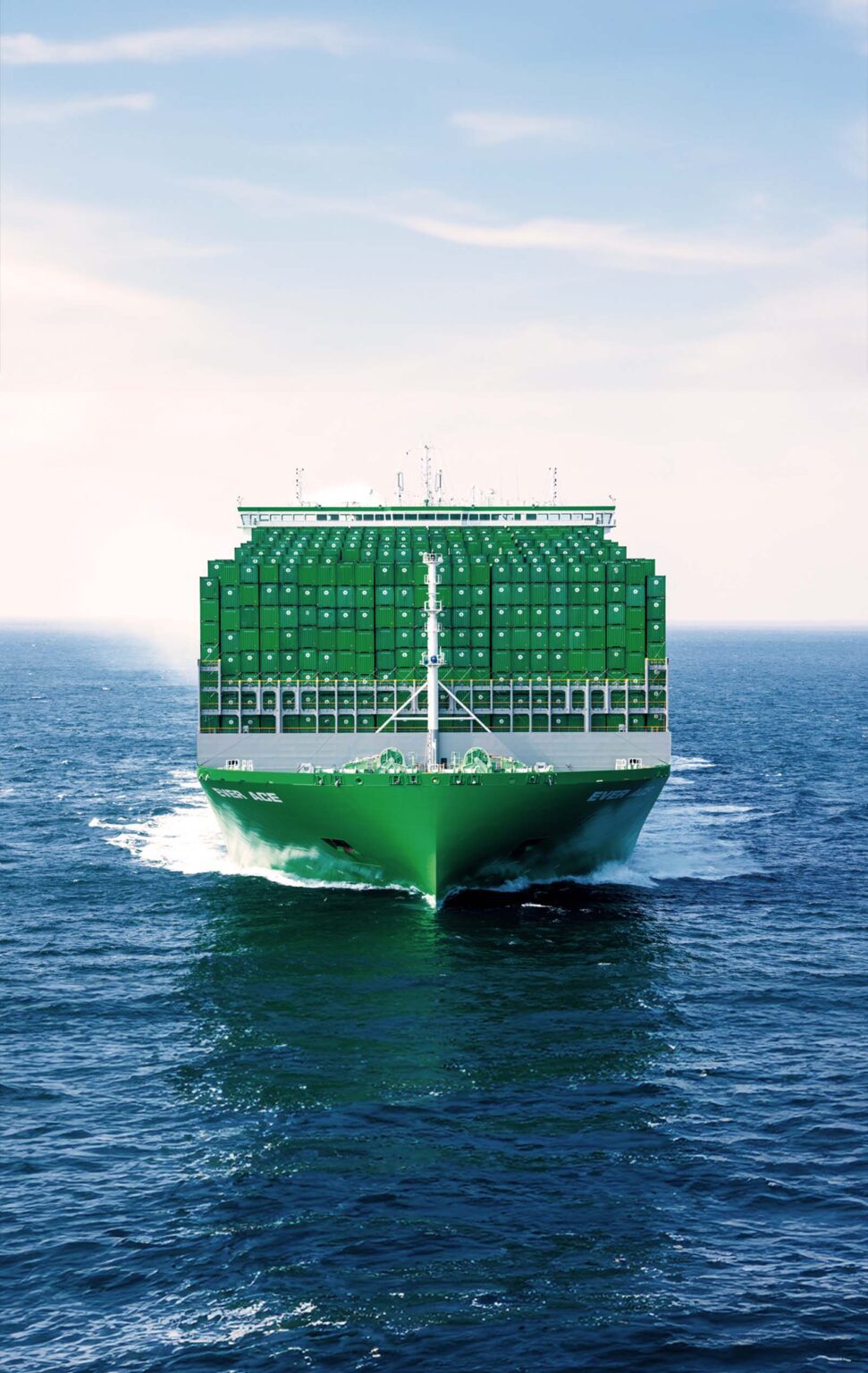 A large green Evegreen cargo ship filled with stacked containers is navigating through blue ocean waters. The sky is clear with a few scattered clouds. The ship's bow is cutting through the waves, creating white froth as it moves forward. The containers create a solid wall of green, indicating a full load. The scene conveys a sense of robust maritime commerce and the vastness of oceanic travel.