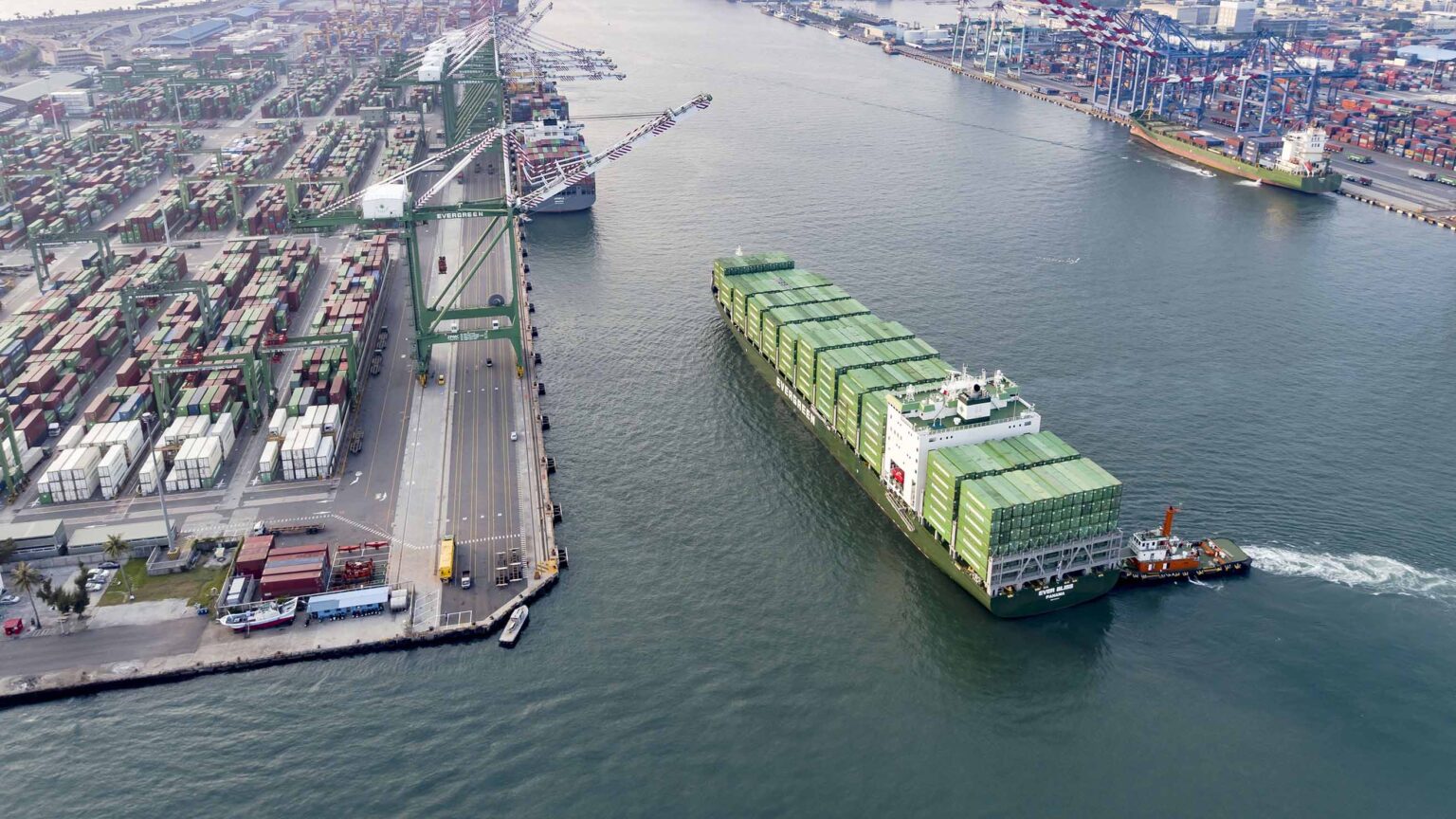 A bird's-eye view of a busy cargo port. Multiple rows of colorful shipping containers are neatly stacked on the dock, and container cranes tower above. A large Evergreen container ship, adorned with rows of green evergreen containers, is maneuvering out of the port with the help of a small tugboat. The water is calm, and the skies are overcast, casting a soft light on the industrial scene.